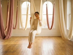 Beginner zweeft zachtjes in een crèmekleurige aerial yoga hangmat in een zonnig Amsterdams studio met houten vloer.