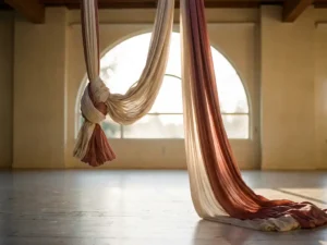 Zijden aerial yoga hangmat in twee draperingen: strak gevouwen en zacht golvend op houten vloer, warm middaglicht, ivoor en roze tinten.