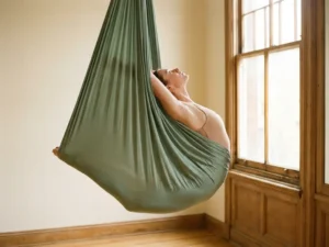 Danser in diepe rugbuiging hangend in een salie groene luchthangmat, borst open naar zacht ochtendlicht vanuit studiovenster.